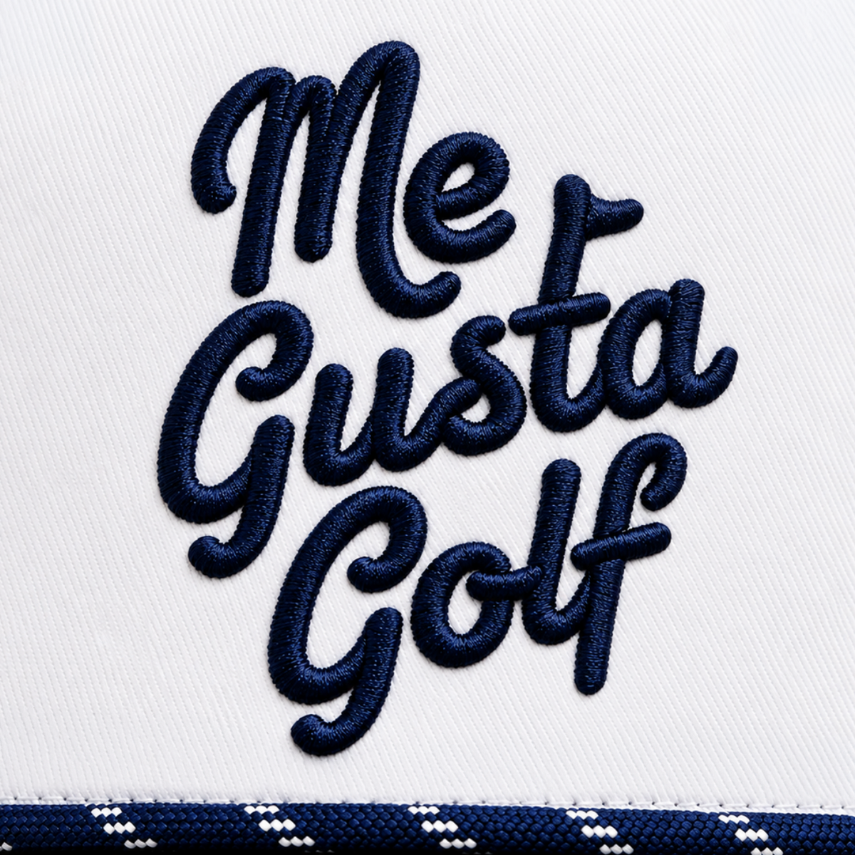 Close-up of a white embroidered cap with the phrase 'Me Gusta Golf' in dark navy script across the front, plus a navy and white patterned brim edge visible at the bottom.