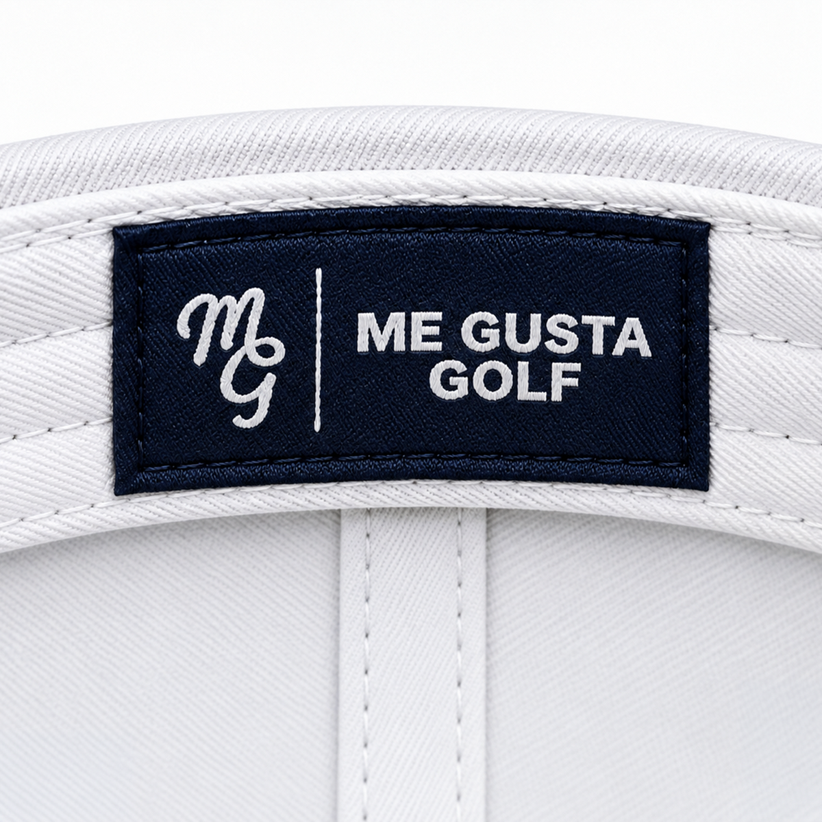 Close-up of a white golf cap interior label featuring a navy blue rectangular patch with white embroidery reading 'ME GUSTA GOLF' and a stylized 'MGG' logo.