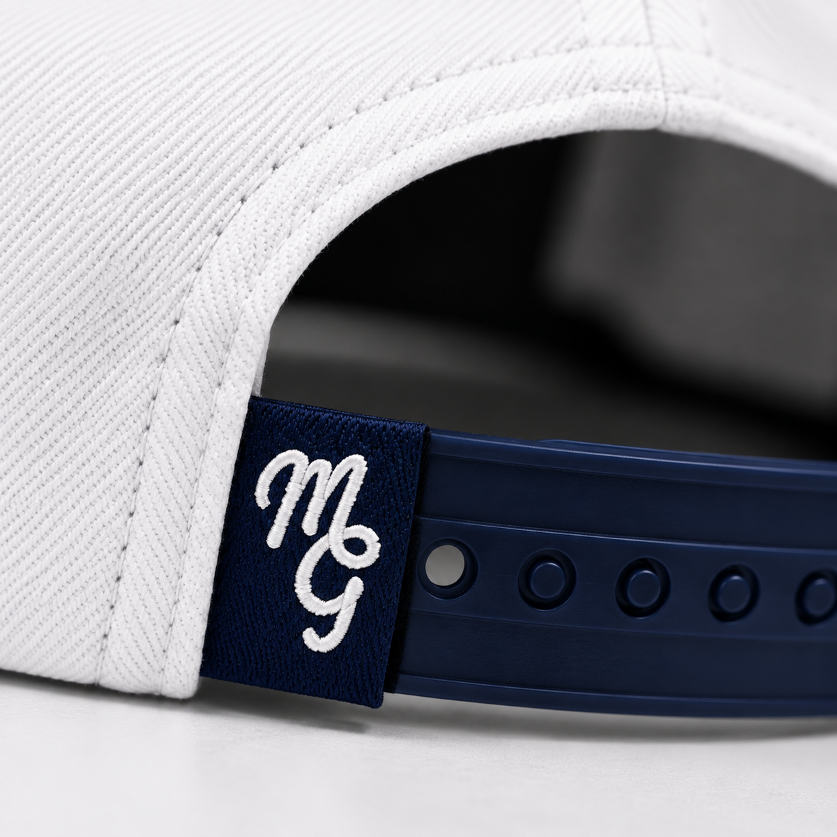 Close-up of a white structured cap showing the back strap closure with a navy blue adjustable band and a matching navy patch embroidered with a white stylized 'MGG' logo.