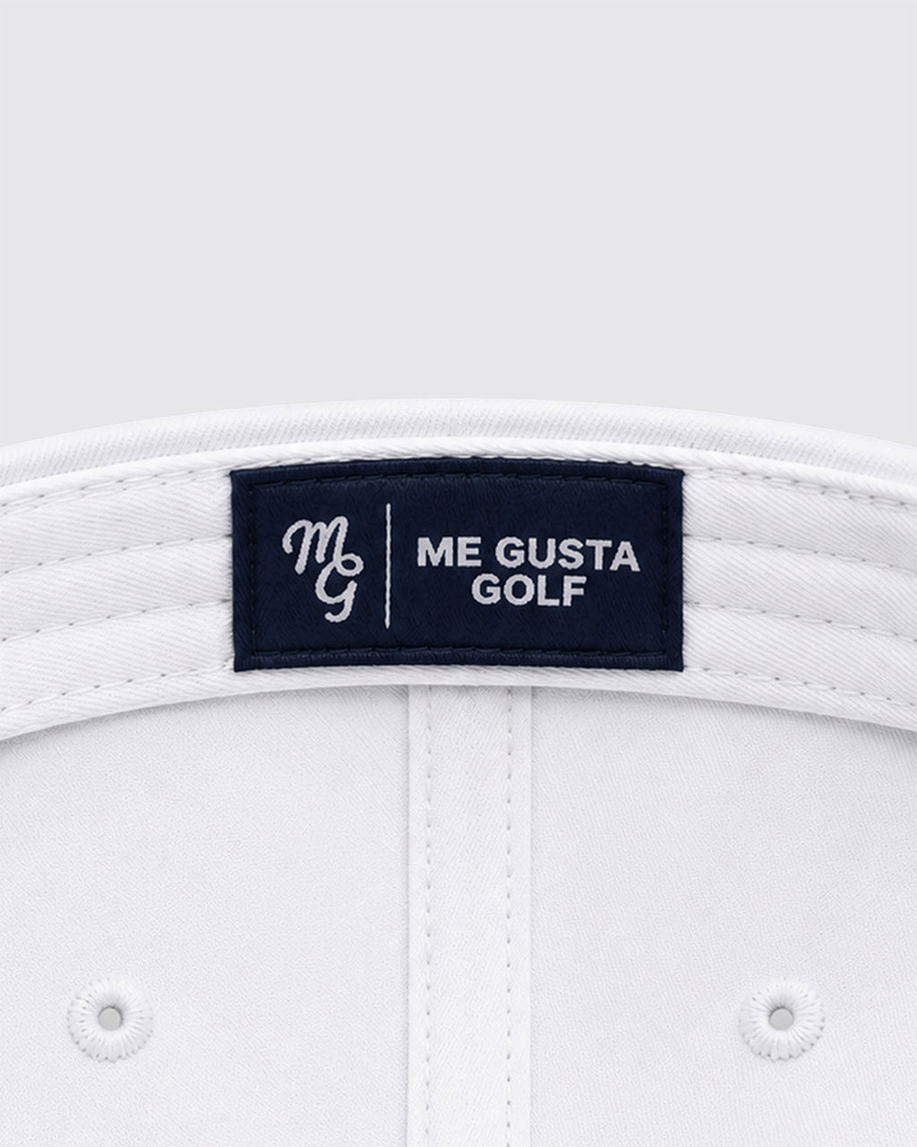Close-up of the inside crown of a white golf hat, showing a dark navy brand label that reads 'ME GUSTA GOLF' with a white embroidered monogram.