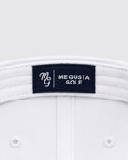 Close-up of the inside crown of a white golf hat, showing a dark navy brand label that reads 'ME GUSTA GOLF' with a white embroidered monogram.