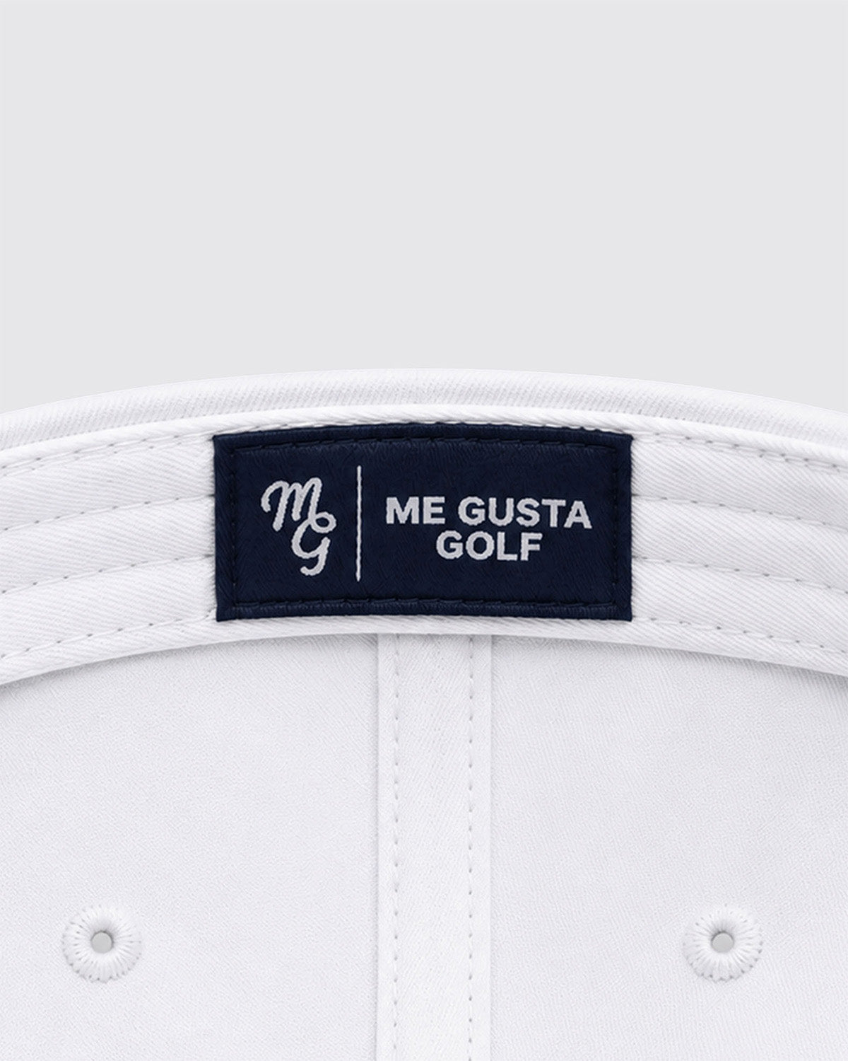 Close-up of the inside crown of a white golf hat, showing a dark navy brand label that reads 'ME GUSTA GOLF' with a white embroidered monogram.
